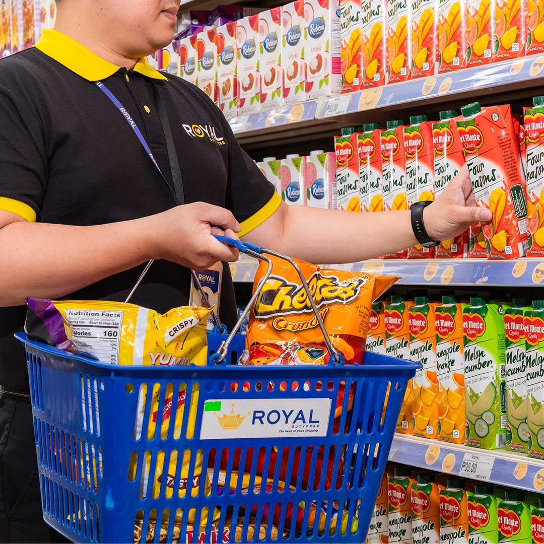 A Royal Personal Shopper in a grocery aisle wearing a black and yellow Royal uniform, holding a blue basket filled with snacks and buying a juice box. 